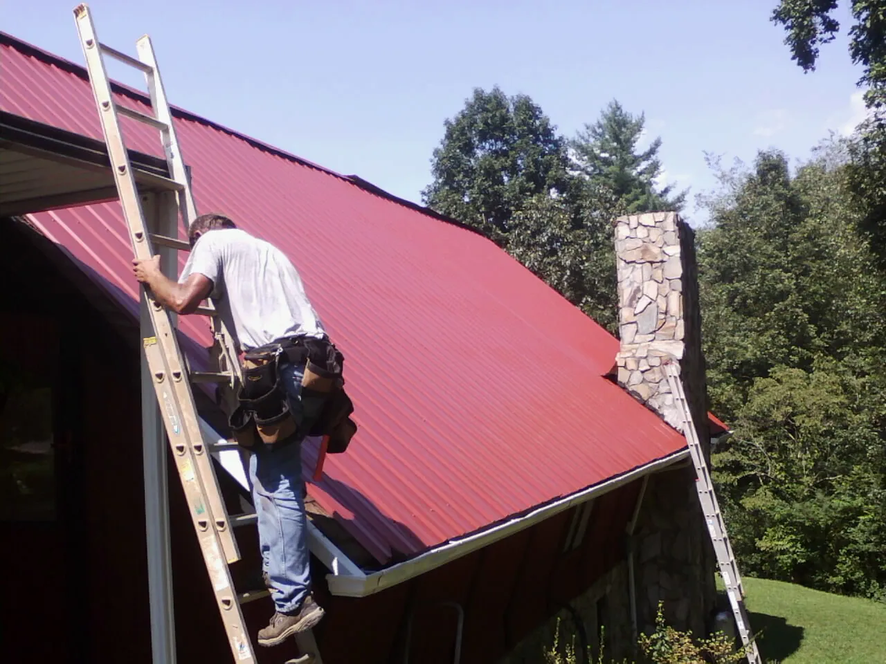 Roofer on a ladder beside a red metal roof and stone chimney — Soffit & Fascia Repair by Roofing Brothers in Pine Hill